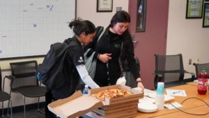 Two women standing at a table with a pizza box in front of them.