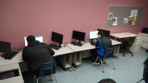 Two individuals working at computers in a room with pink walls and multiple unoccupied workstations.