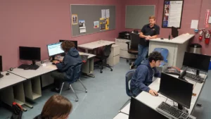 Students working on computers in a classroom with an instructor standing by.