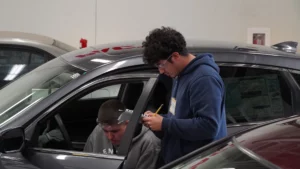 Two individuals inspecting a car, one inside taking notes and the other observing through the window.