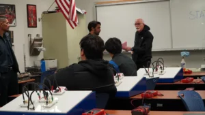 Adult students engaged in an electronics class with an instructor discussing a topic at the front of the room.