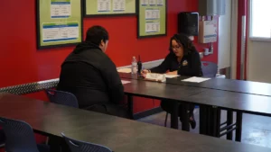 Two individuals engaged in a conversation at a table in a room with informational posters on the wall.