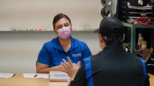 A person wearing a face mask sitting at a desk during a conversation with another individual.