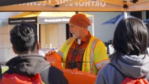 A worker in a high-visibility vest and beanie speaking to two individuals at an outdoor event or workshop.