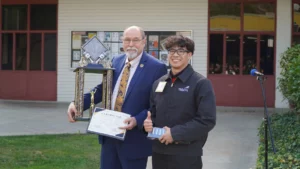 A man in a suit presenting a certificate and trophy to a smiling young recipient at an outdoor award ceremony.