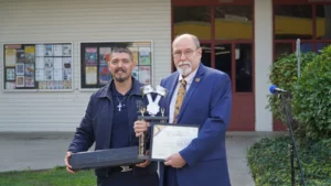 Two men holding a trophy and a certificate of recognition while standing outdoors.