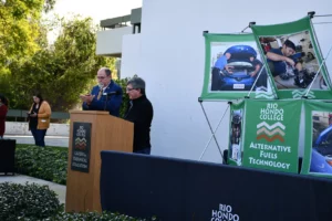 Two individuals at a podium during an outdoor event with banners related to alternative fuels technology in the background.