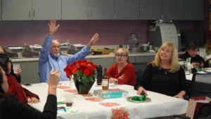 A group of adults gathered in a kitchen for an event, with one man raising both hands enthusiastically.