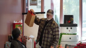 An older man in a hat and plaid jacket holds up a paper bag while standing in a room decorated for christmas.
