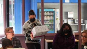 A person wearing a mask carrying a gift box in a room with other people sitting at tables.