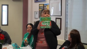 A woman standing and holding up a holiday-themed gift with a smile, while others sit around her indoors.