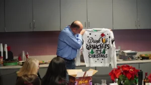 Man holding up a holiday sweater with the text "drink up grinches it's christmas" in a room with people and food around.
