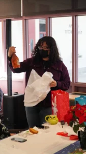 A person standing indoors wearing a mask, holding a gift bag in one hand and a bottle in the other.