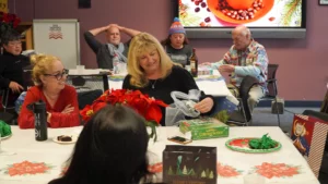 A group of people enjoying a social gathering, with one woman opening a gift while others engage in conversation around a decorated table.