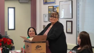 A woman standing and holding a box with the label "bathroom tissue starter kit" while smiling at another seated woman who is also smiling.