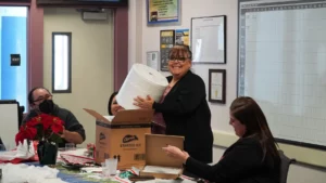 A smiling woman holding a large bag stands next to a cardboard box in an office setting while others look on.