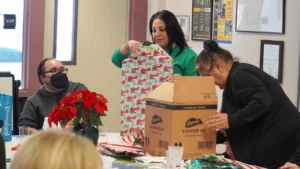 Two individuals exchanging gifts at a holiday event while another looks on.