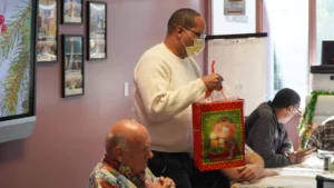 A person wearing a mask stands holding a gift bag in a room where others are seated.