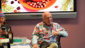 An elderly man in a wheelchair smiling at a social gathering, with a woman beside him and a food-themed picture in the background.