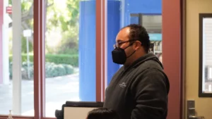 Man wearing a mask standing indoors near a window.