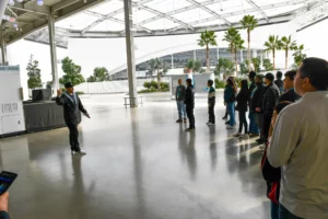 A group of people listening to a speaker in a spacious indoor area with palm trees visible through the glass exterior.