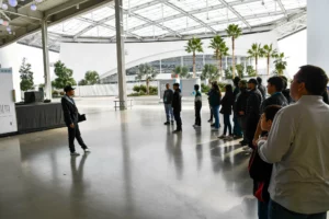 A tour guide addressing a group of visitors inside a large, modern building with a glass ceiling and palm trees in the background.