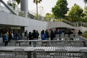 People queueing at a food stand in an outdoor plaza with seating area in the foreground.