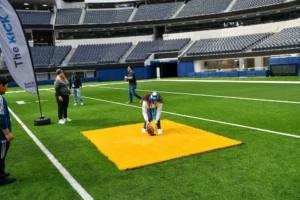 Person in a mascot costume kneeling on a yellow mat at an indoor stadium field with onlookers standing by.