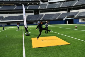 Individual kicking a football on a stadium field with others present.