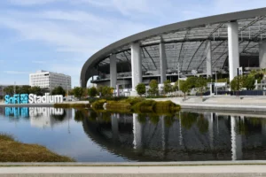 Sofi stadium in inglewood, california, reflects on the surrounding water feature under a cloudy sky.