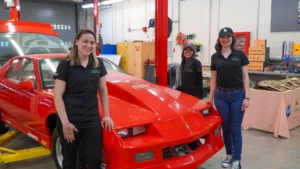 Three women wearing team shirts standing with a red race car in a garage workshop.