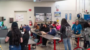 Group of people socializing in a workshop or classroom environment with tables and equipment in the background.