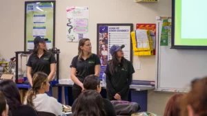 Three individuals in uniform attentively participating in a meeting or training session with informational posters and a presentation in the background.