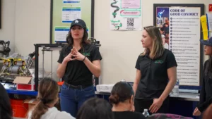 Two women in black shirts presenting in front of a group, with one gesturing while speaking and the other listening attentively.