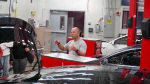 A person explaining something in an automotive workshop with cars and equipment around.