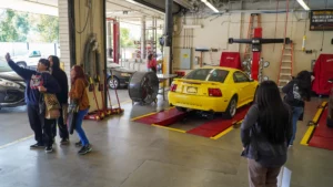 Students observe a car maintenance training session in an automotive workshop.