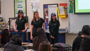 Three women in black polo shirts stand at the front of a classroom during a presentation.