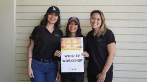 Three smiling individuals holding a sign for a wing-ev workshop.