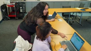 Two women collaborating on laptops in a classroom setting.