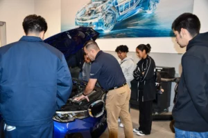 A group of students observe a mechanic demonstrating engine repairs on a blue car in a workshop.