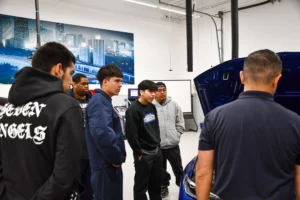 A group of students listens attentively to an instructor in a car mechanics workshop, standing around an open car hood.