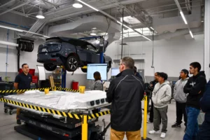 A group of people attentively watch an instructor in an automotive workshop, with a car raised on a lift above them.