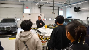 A group of students listens to an instructor in an automotive workshop surrounded by various car parts and two luxury cars.