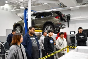 Automotive students observing a car elevated on a lift in a training workshop.