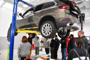 A group of people observes a car being lifted on a yellow hydraulic lift in a workshop.