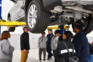 A group of students and an instructor examining a car lifted on a hoist during an automotive training class.