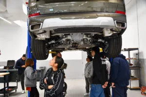 Group of people observing a car raised on a lift in an automotive workshop.
