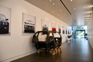 A car shell on casters displayed in a gallery hallway with framed images of various car models hanging on the walls.