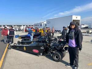 A person standing in front of drag racing cars in a pit area with several people and trailers in the background.