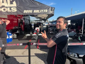 Man gesturing with a smile in front of the doug kalitta racing team tent at a motorsport event.